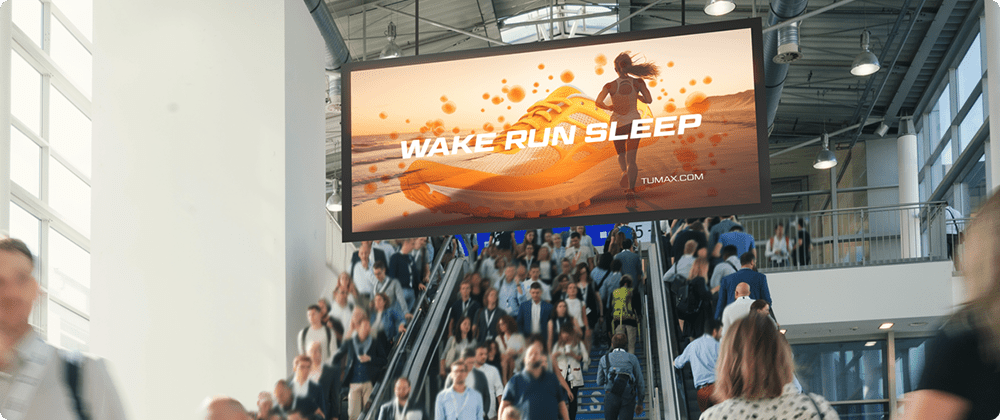 Digital out-of-home display showing a running-themed ad above a crowded transit concourse with heavy foot traffic.