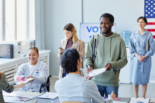 Man casting his ballot at a polling station while two women wait in line, one looking at her phone.